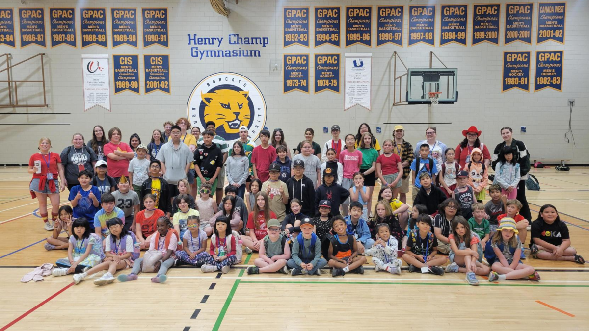 A group photo of dozens of children sitting in a gynmasium. Some older teenagers and young adults wear T-shirts that say Mini U on them.