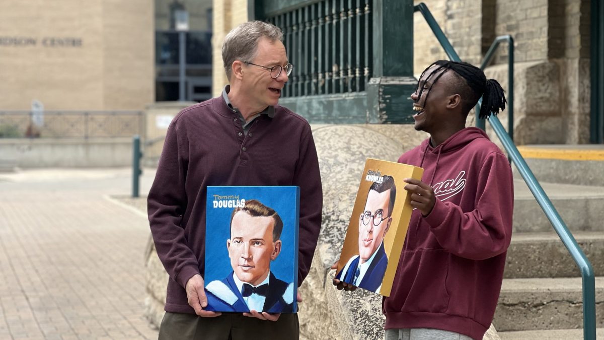 A man and a university-aged student look at each other while laughing. They are carrying paintings of two other men.