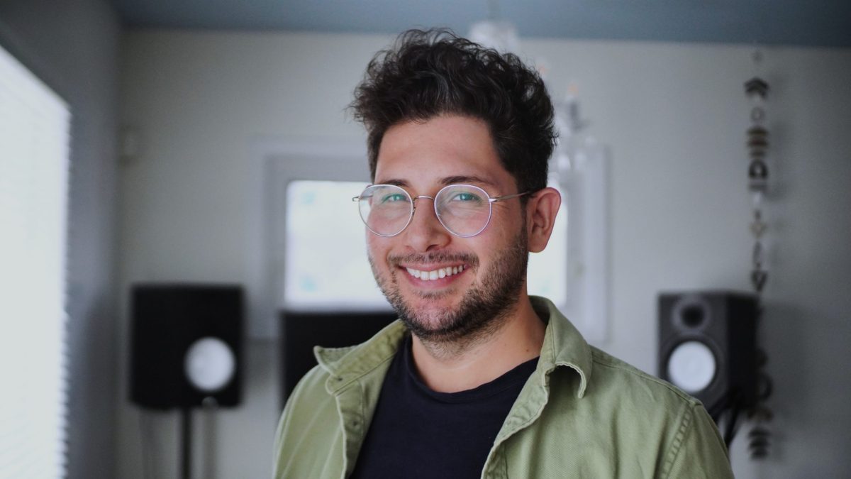 A man smiles in a white room. Audio speakers can be seen behind him