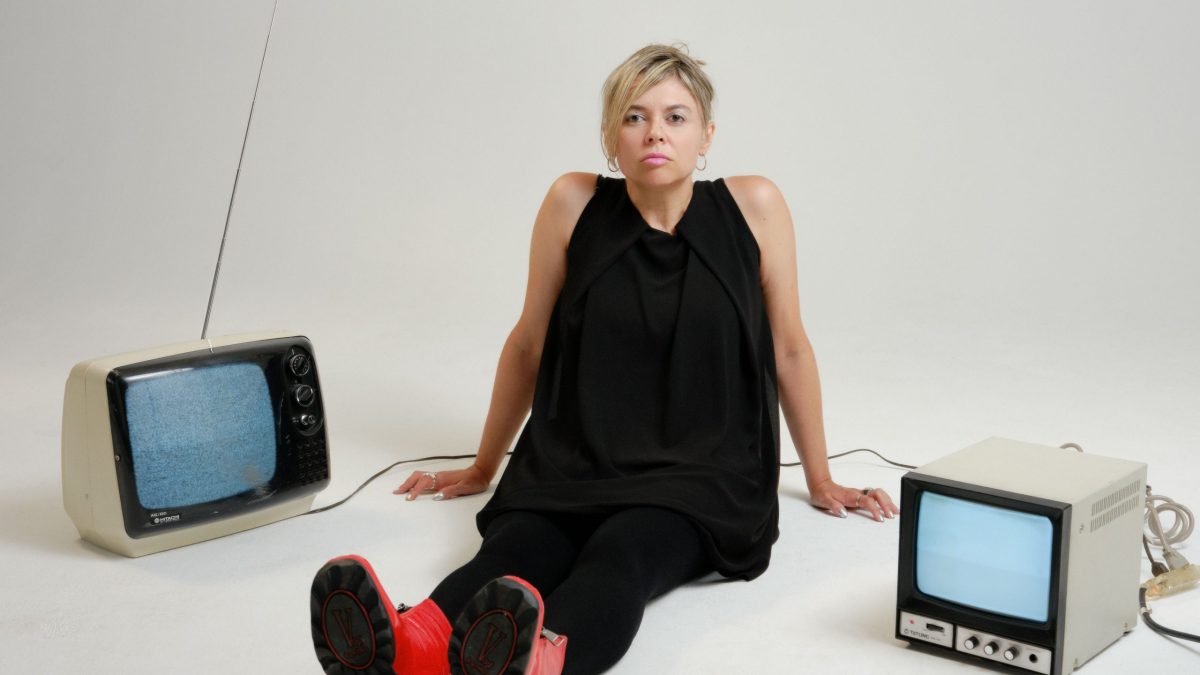 A woman sits in a white room between an old television and an old computer monitor