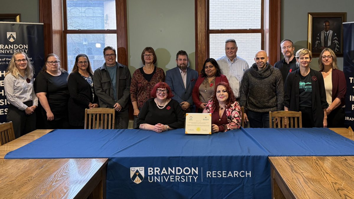 Two women sit at a table with a table cloth saying "Brandon University Research." A large group of people stand behind them. A certificate is propped on the desk.