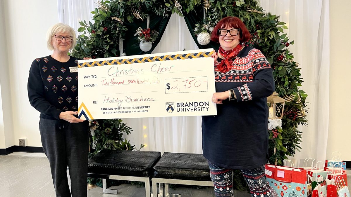 Two women stand in front of holiday garland with an oversized promotional cheque that reads "$2,750" made out to Christmas Cheer