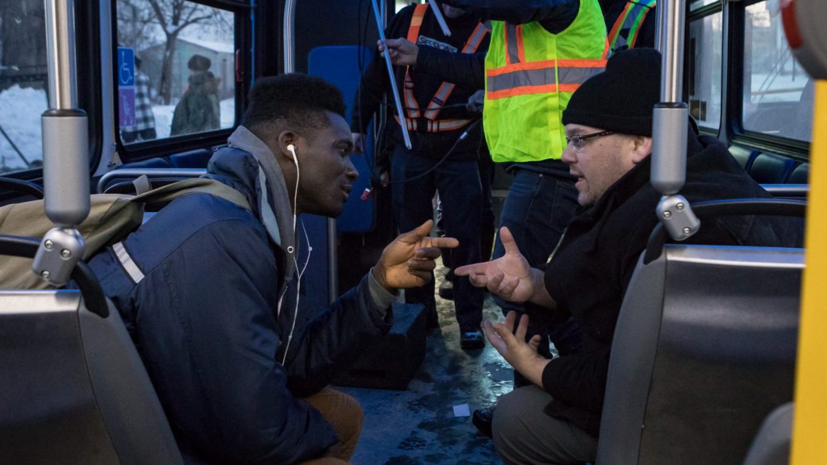 Two men speak across the aisle while seated on a bus