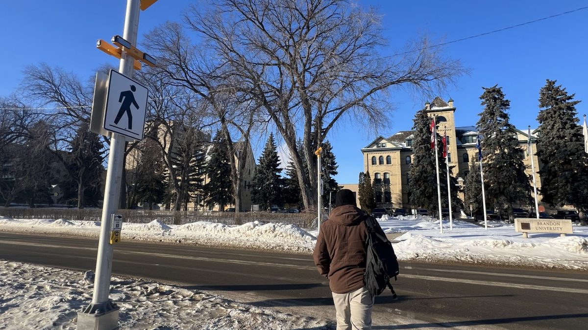 A person walks across a wide road in front of a university. There is a pedestrian crossing sign and warning lights.