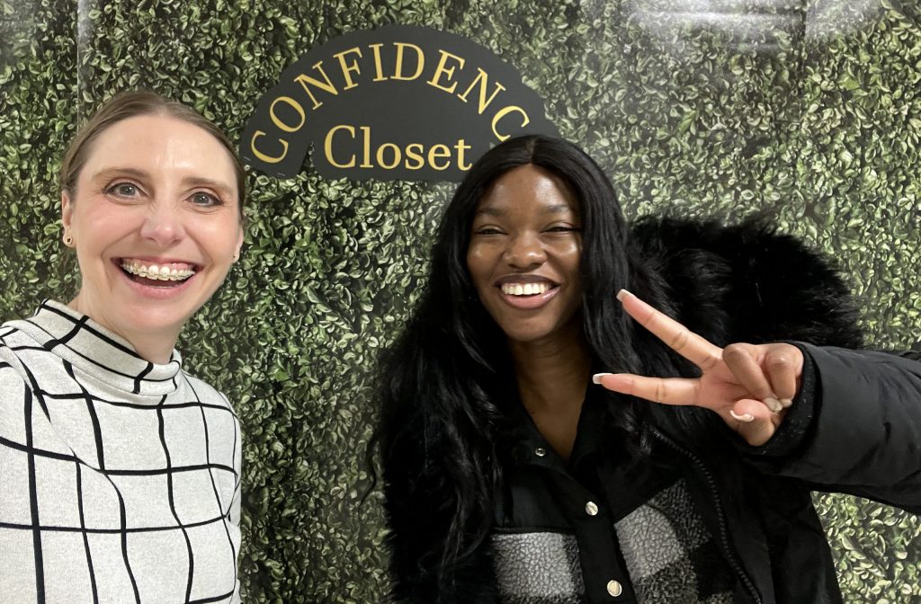 Two women smile and pose in front of a decorative wall with the words "Confidence Closet" on them.