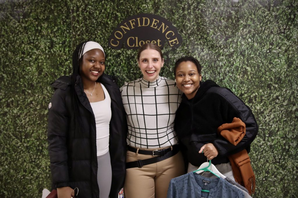 Three women smile and pose in front of a decorative wall with the words "Confidence Closet" on them.
