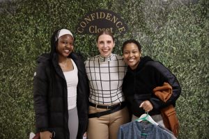 Three women smile and pose in front of a decorative wall with the words "Confidence Closet" on them.
