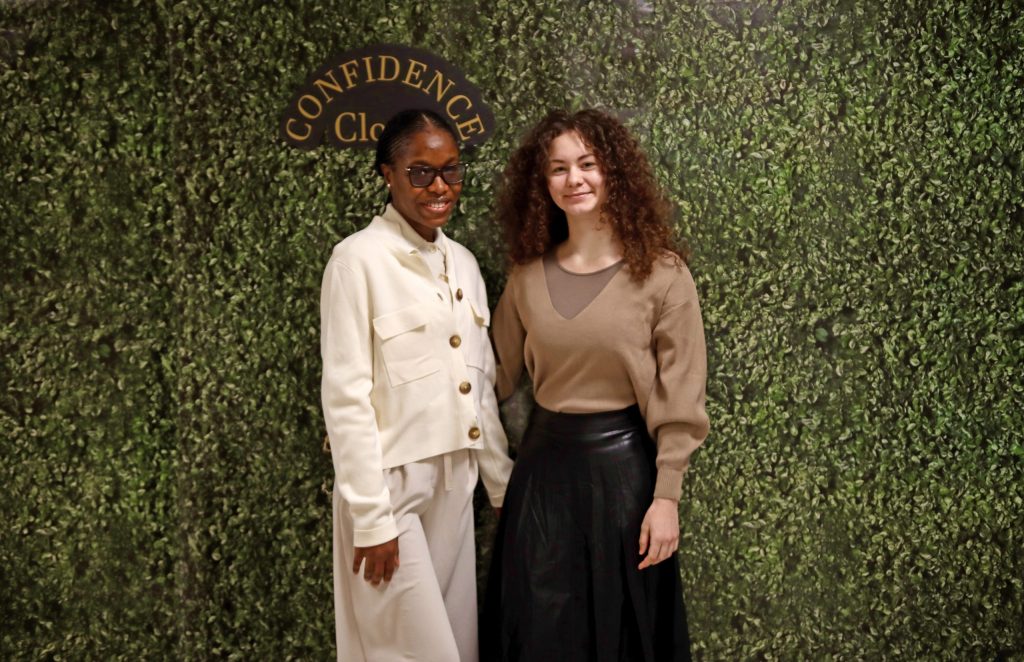 Two women stand in front of a decorative wall with the words "Confidence Closet" on them.