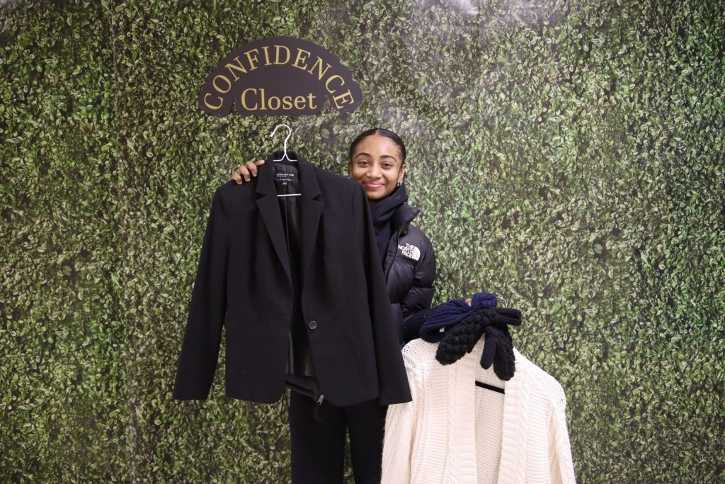 A woman holds up pieces of clothing on hangers in front of a decorative wall with the words "Confidence Closet" on them.