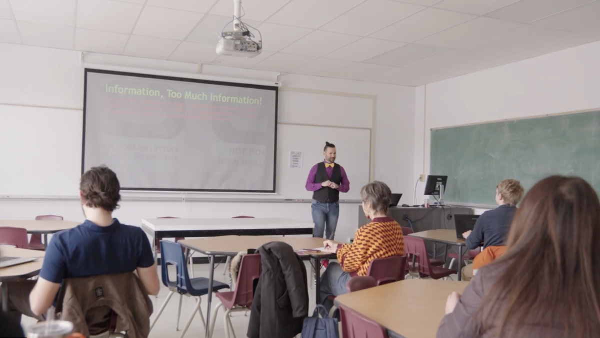 A person stands and speaks in the front of a classroom. The backs of several attendees can be seen.