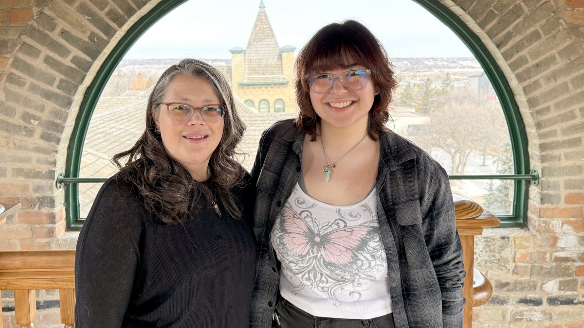 Two women smile in front of a semicircular window. A spire on an old window can be seen between them in the background