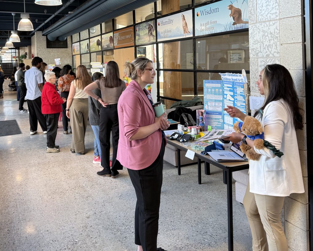 People gather around a lineup of desks with display boards on them in a hallway.