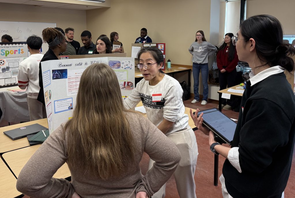 A woman speaks to a group of people gathered around a display board.