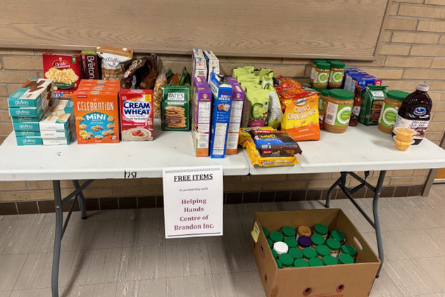 A table with several packaged food items on top. A box with more jars of food is below the table