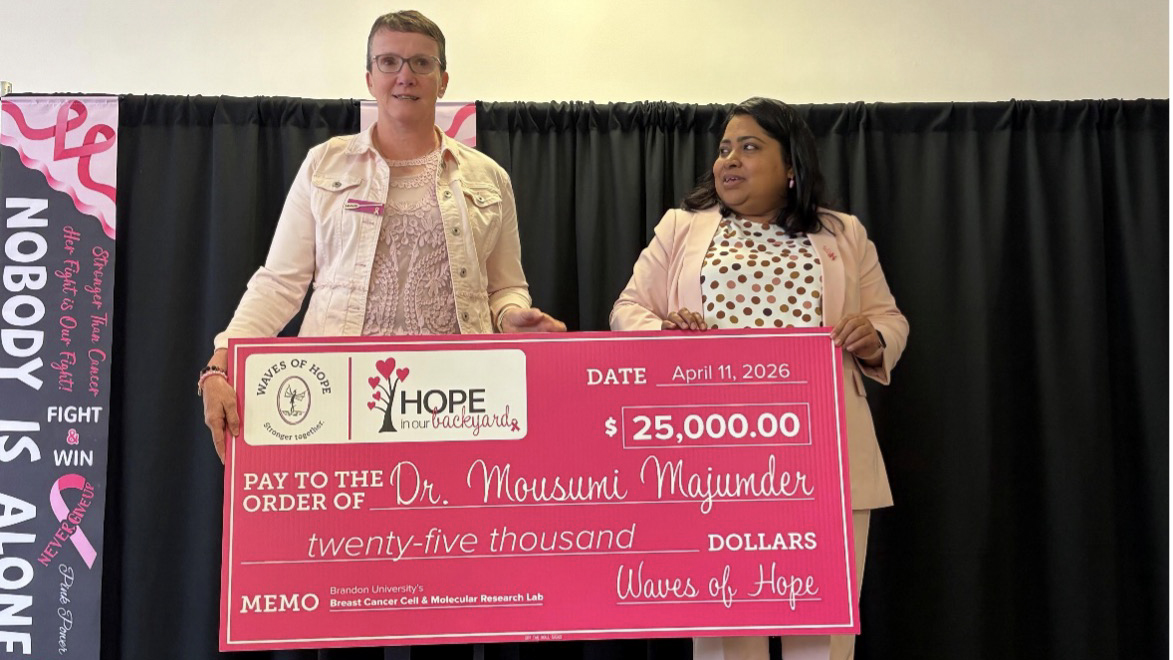 Two women stand on a stage with an oversized novelty cheque. It is pink.