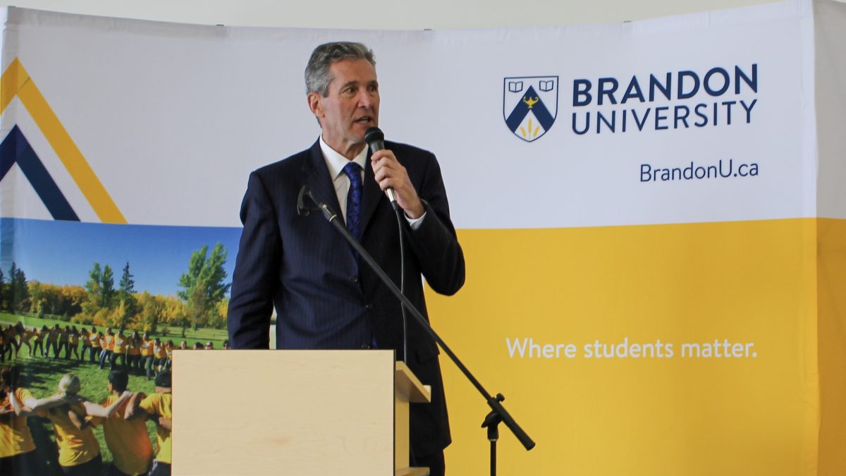 A tall man speaks at a lectern in front of a Brandon University backdrop.