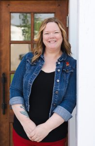 A woman leans on a wall and smiles while posing in front of a wooden door
