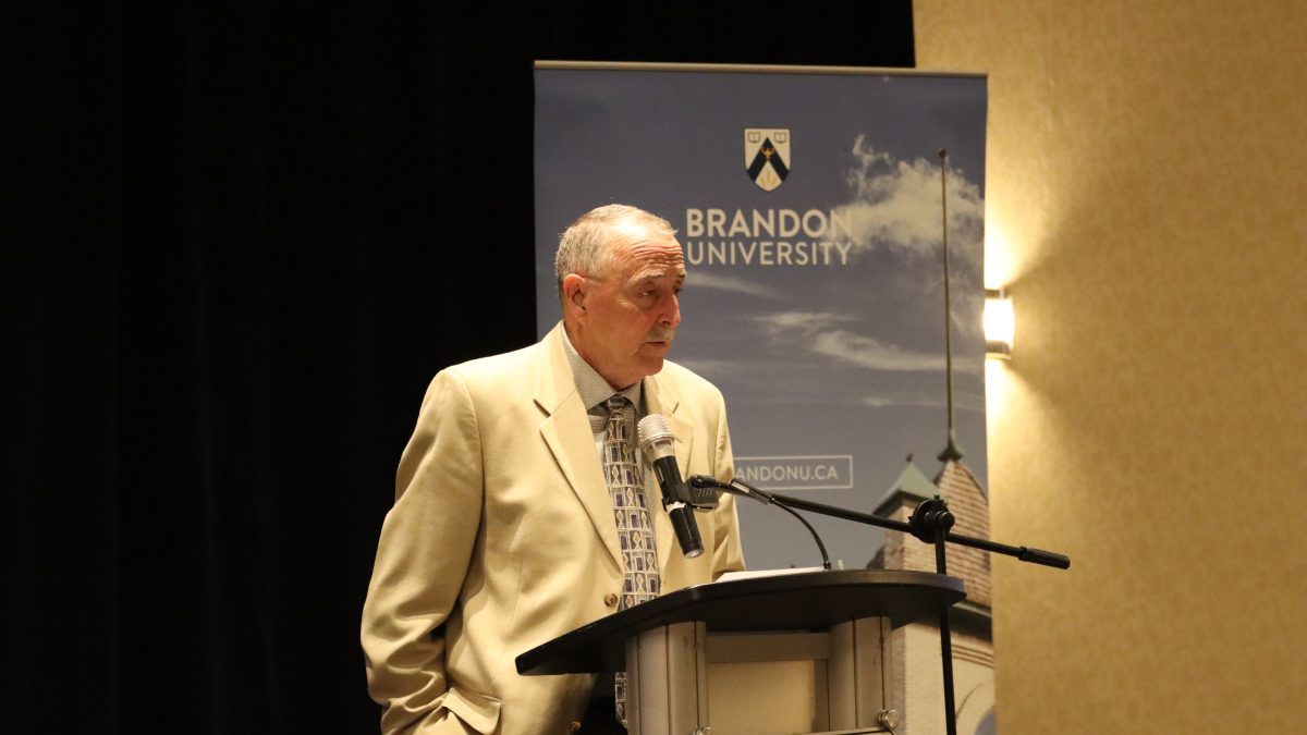 A man speaks at a podium, with a Brandon University banner behind him.
