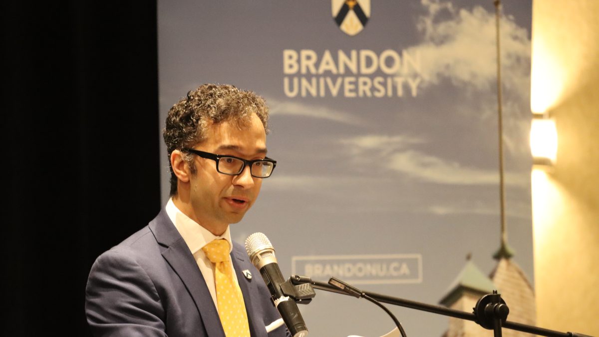 A man in a suit stands at a podium with a Brandon University banner behind him.