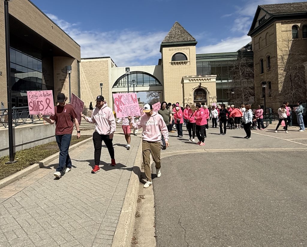 A large group of people walk though a courtyard. One of them is carrying a sign that says "Early Detection Saves Lives," while another his holding a sign that says "Know More"