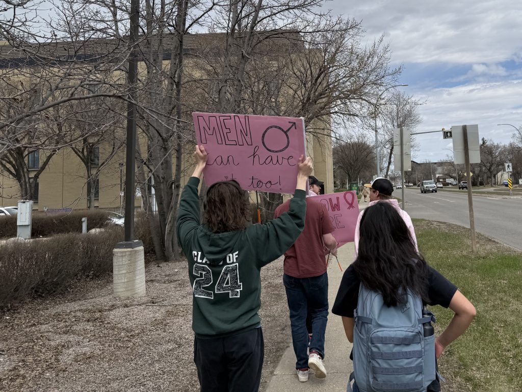 A group of people walk along a street. One of them is holding up a sign that says "Men can have it too"