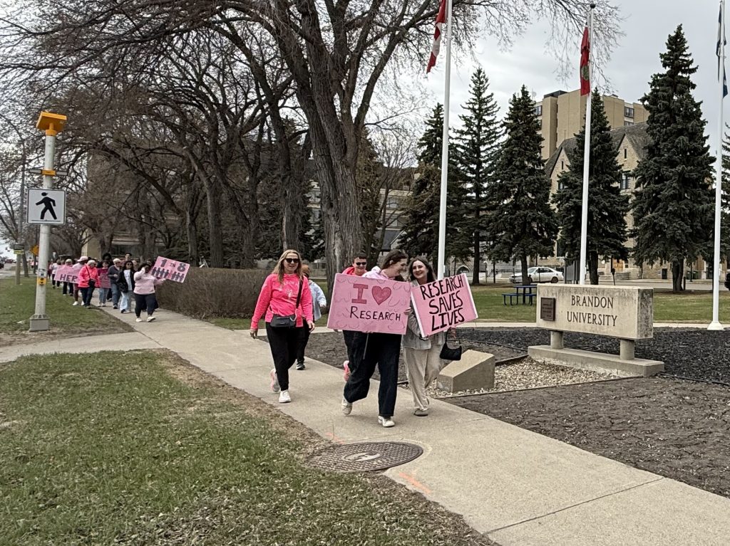 A group of people walk past a Brandon University sign. One person is holding a sign that says "I (heart) research," while another has a sign that says "Research Saves Lives"