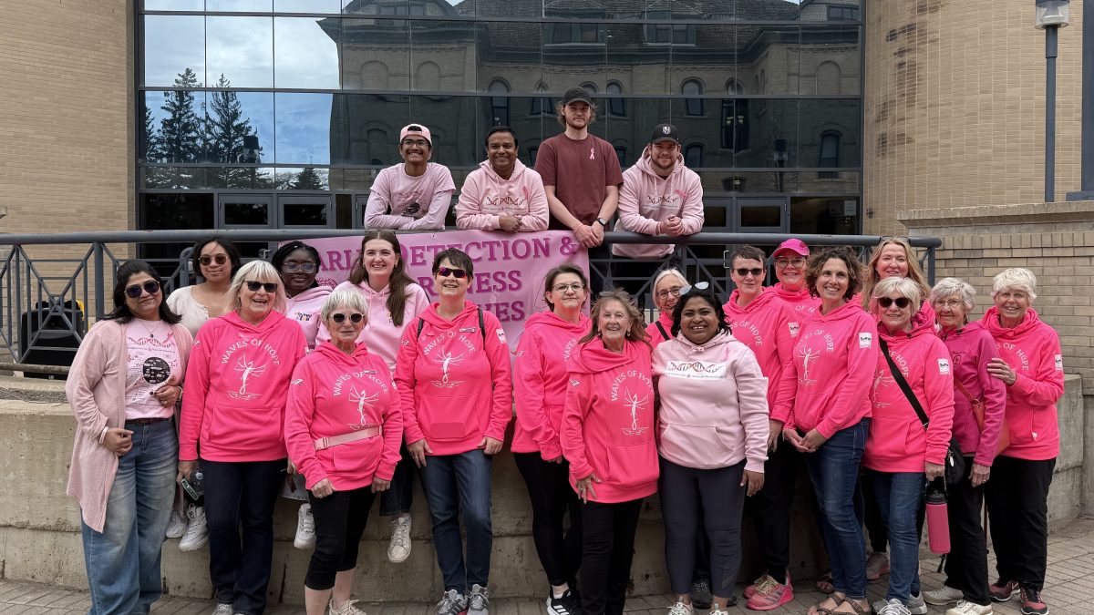 A large group of people wearing pink shirts pose for a photo. They are in front of windows that reflect the image of a brick building.