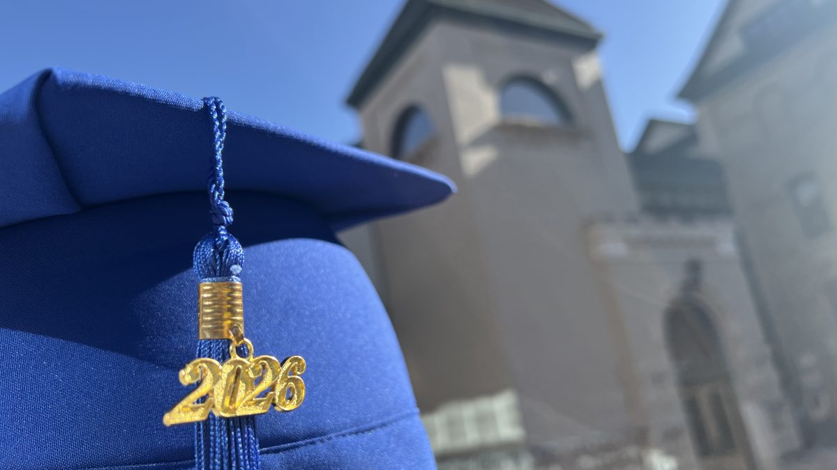 A blue graduation cap is pictured in front of a brick tower. The cap features a "2026" charm