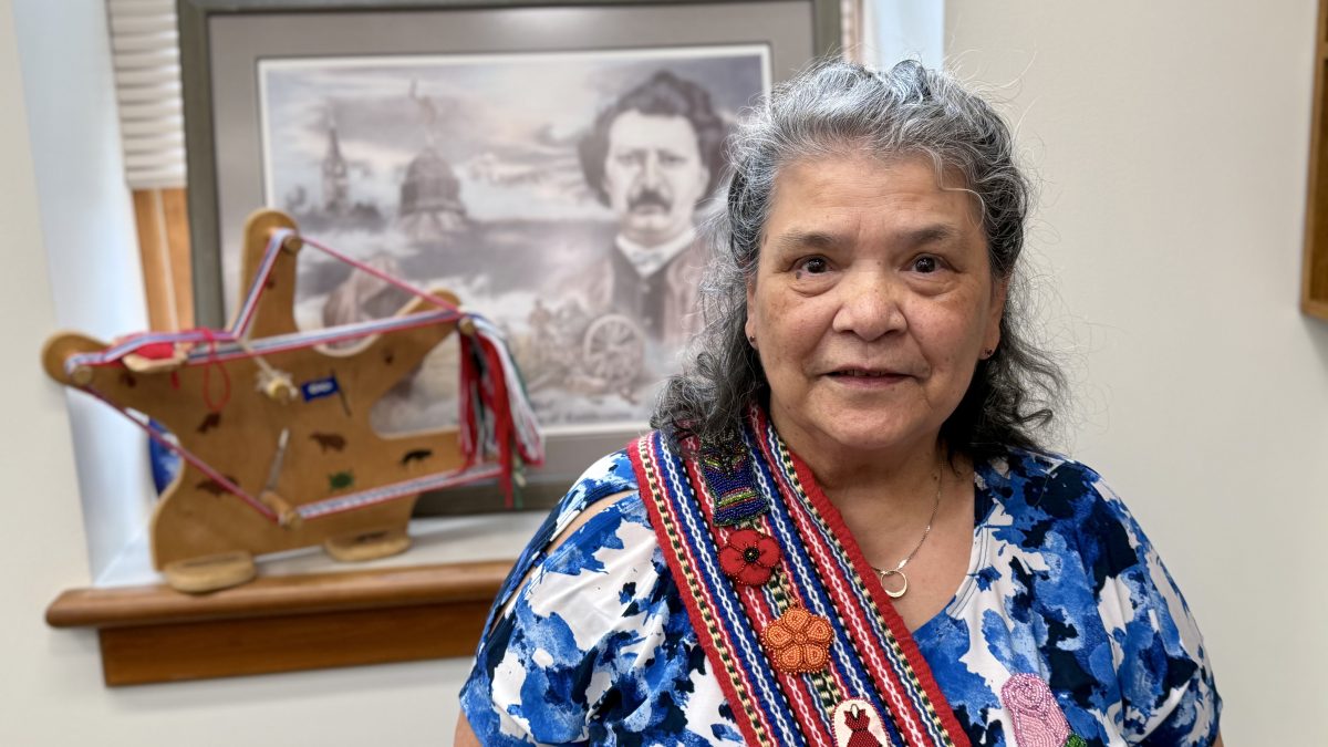 A woman poses wearing a sash decorated with beaded designs. Behind her are a loom with wool on it and a picture of Louis Riel.
