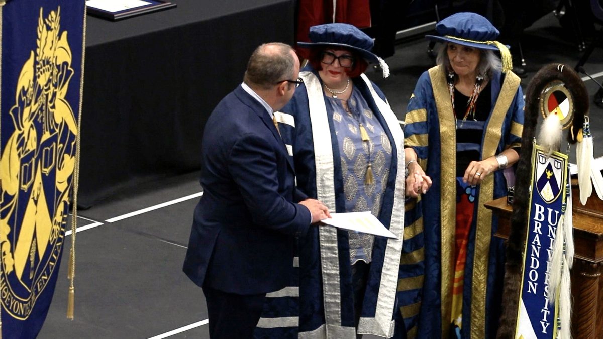 A woman in academic garb stands between a man in a suit and another woman in academic garb.
