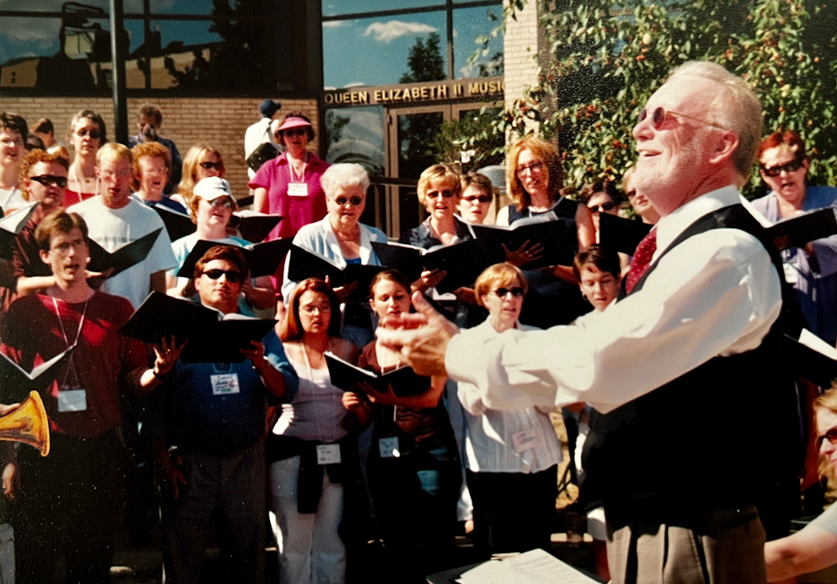 A conductor looks up and smiles as several singers sing.