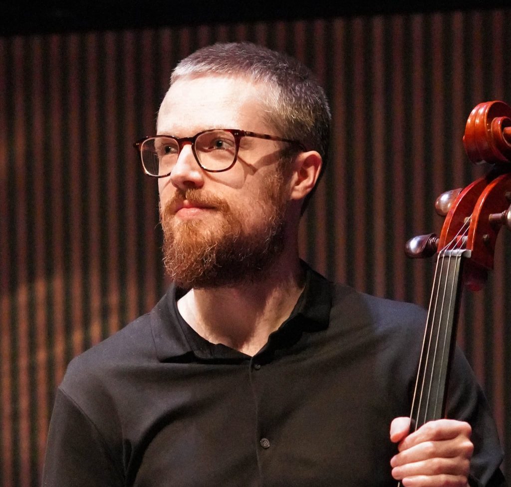 A man looks to the side. He is holding a cello in his hand