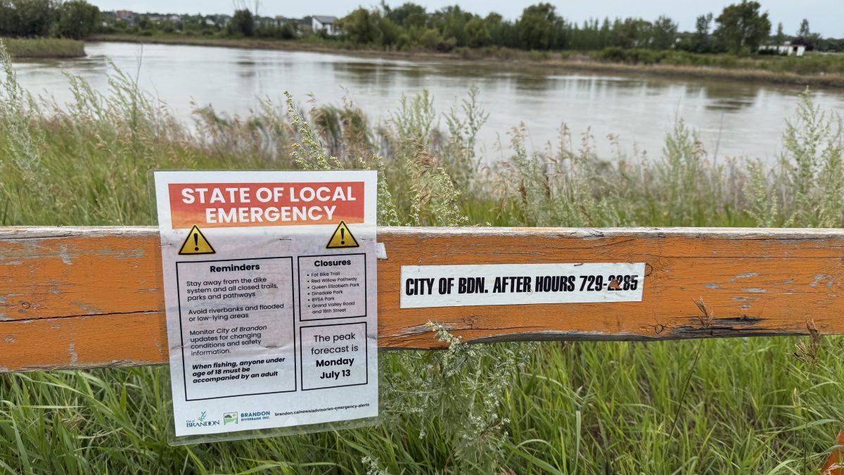 An orange barrier is seen in front of tall grass and a river. A sign saying "State of Local Emergency" is attached to the barrier