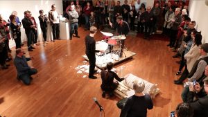 A man sits at a drumset at the centre of a room and hits porcelain discs while surrounded by observers. Another man stands beside the drumset waiting to present him with the next porcelain disc.