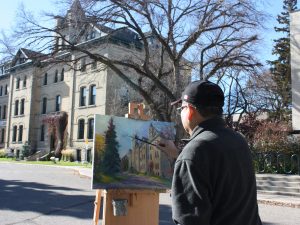 A man stands at an easel painting a university building. The building is seen in the background.