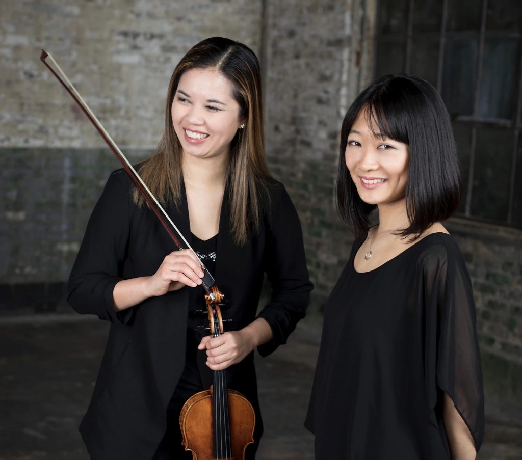 Two women smile. One woman is holding a violin and bow