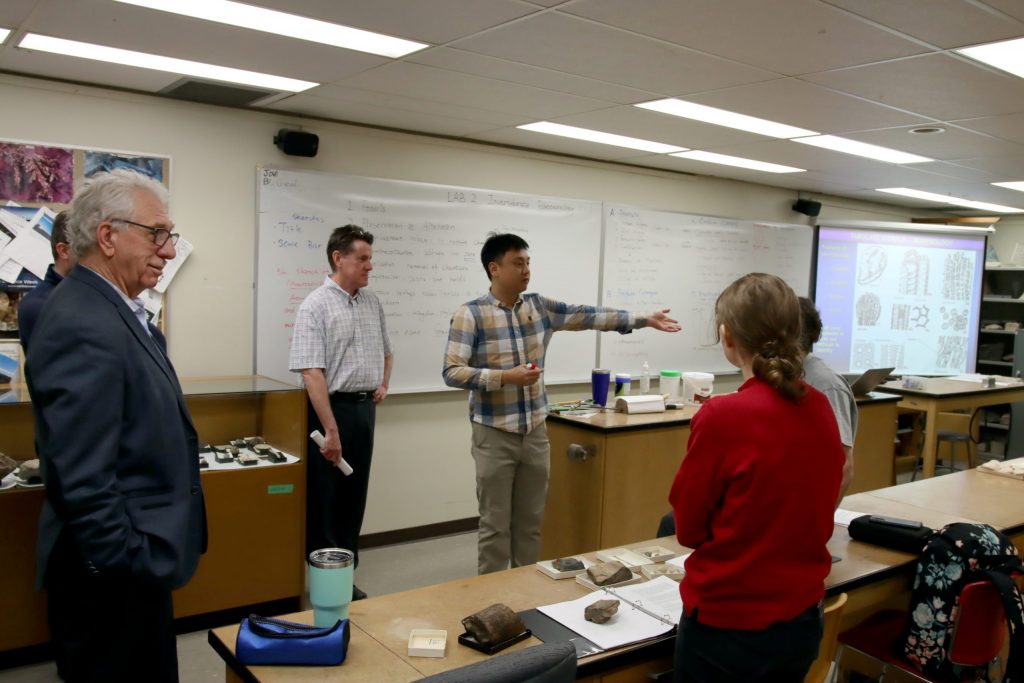 A group of men stand in front of a student in a lab