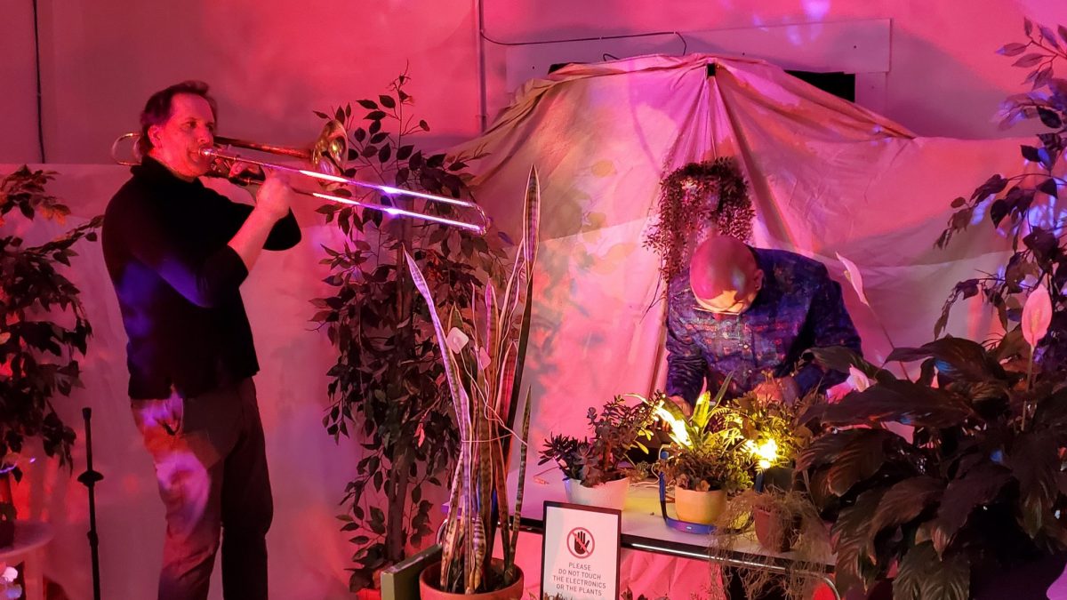A trombone player and an electronic musician perform while surrounded by plants