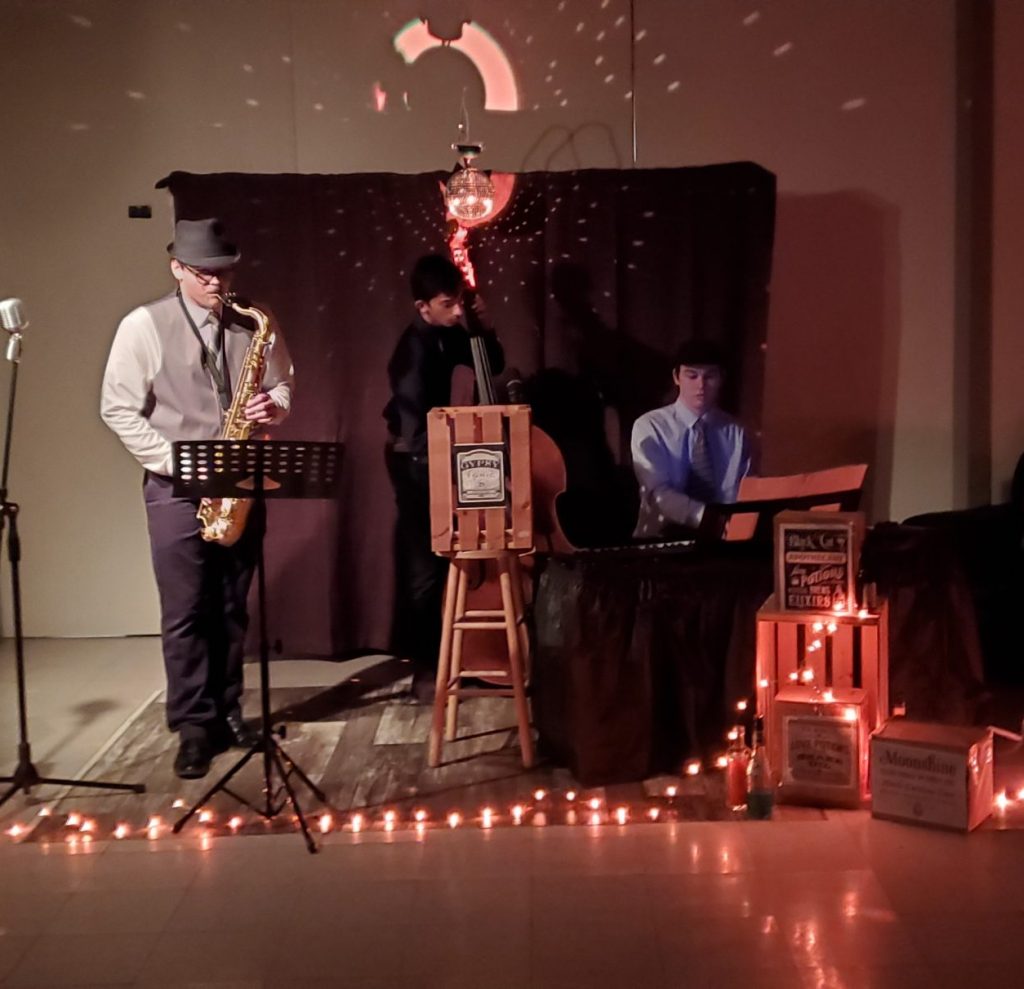 A saxophonist, an upright bassist and a keyboardist play while surrounded by lights