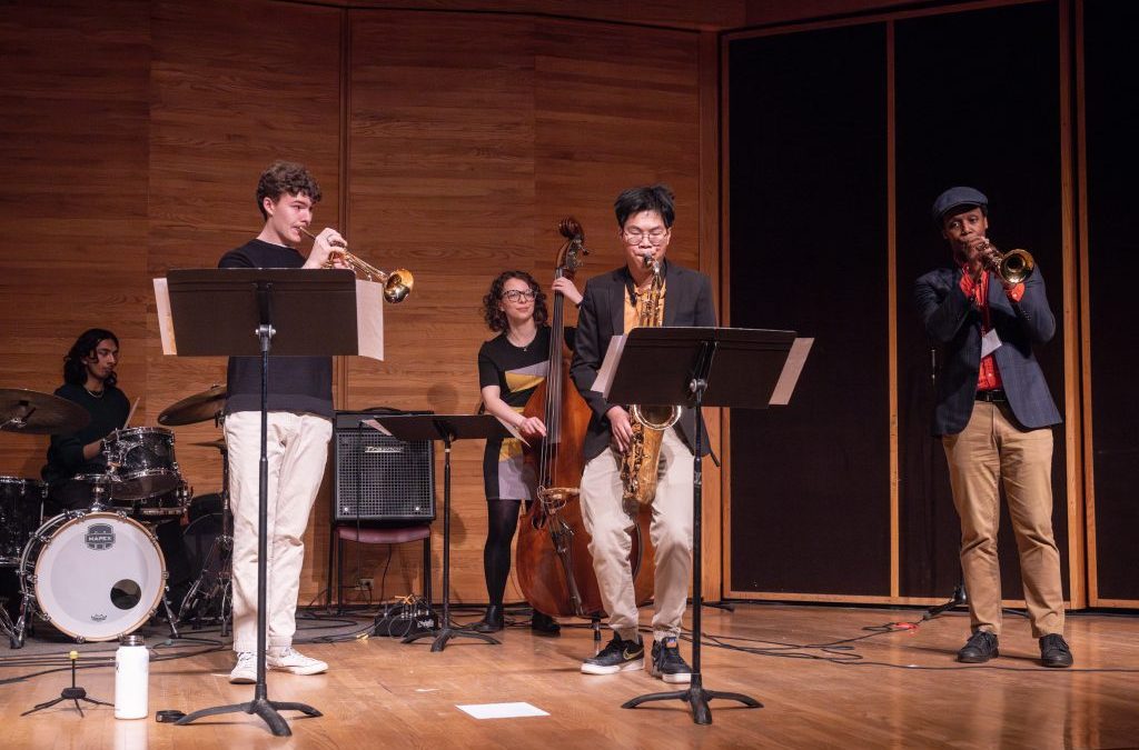 A group of musicians play drums, trumpets, saxophone and an upright bass on a stage