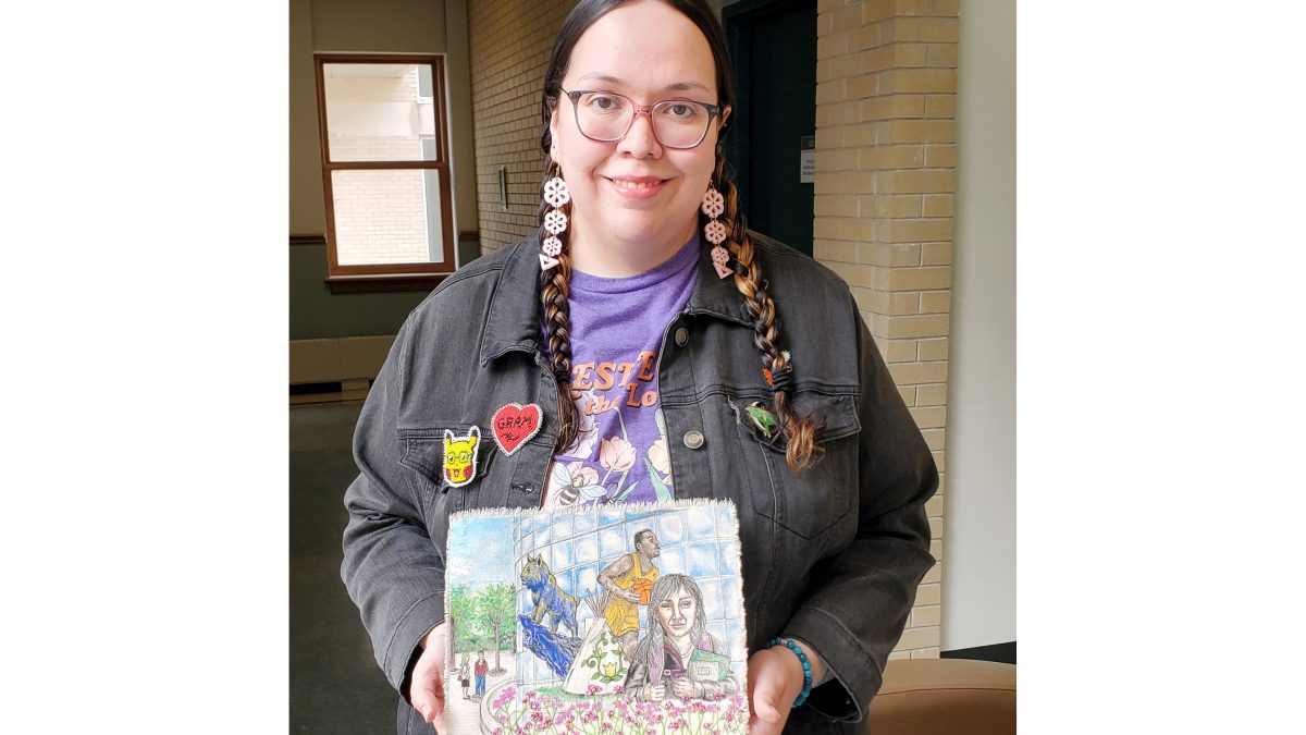 A woman smiles while holding canvas art. The art shows an Indigenous student, a tipi decorated with florals, a basketball player, a bobcat statue and flowers in front of a curved window, with itger students in the background