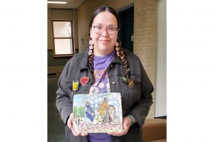 A woman smiles while holding canvas art. The art shows an Indigenous student, a tipi decorated with florals, a basketball player, a bobcat statue and flowers in front of a curved window, with itger students in the background