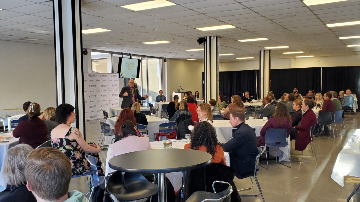 A large group of people sit at tables while a man stands and speaks in front of them.