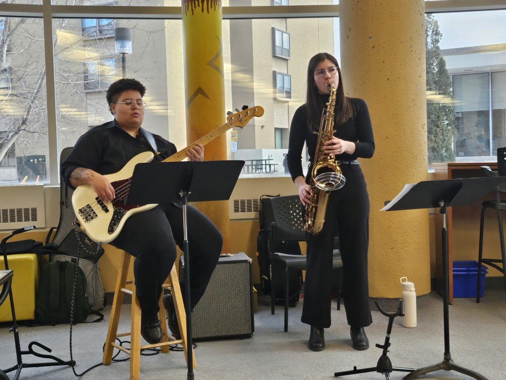 A bass play sits on a stool while playing next to a standing saxophonist