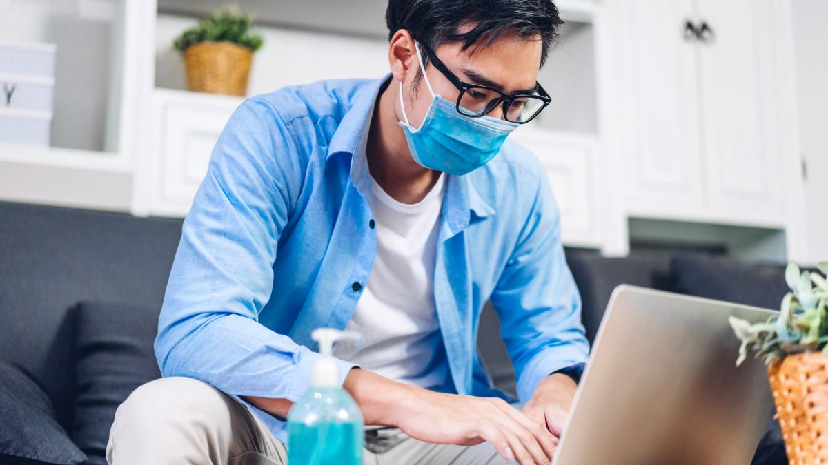 Young asian man using laptop computer working and video conference meeting online chat in quarantine for coronavirus wearing protective mask with social distancing at home.