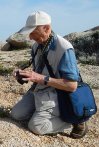 Al Rogosin kneels and photographs a plant while in a rocky area.