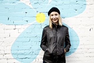 A woman smiles while standing with her hands in a picket. Behind her is a brick wall with a painting of a blue flower.