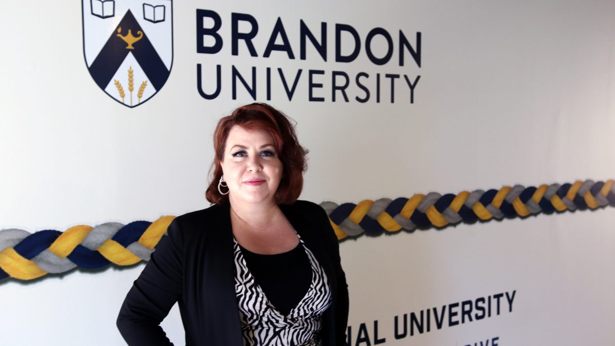 A woman stands in front of a wall with a Brandon University logo