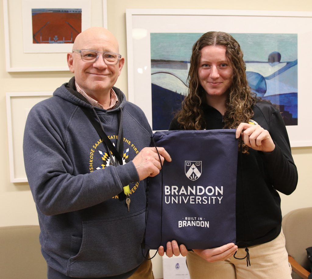 A man and a woman smile while holding a Brandon University bag between them.