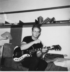 Vintage, black-and-white photo of a man sitting on a chair playing an electric guitar